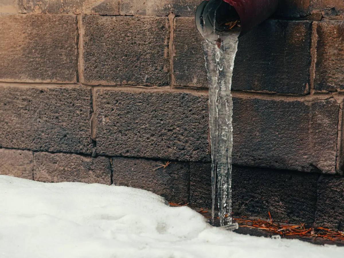 Water pouring from a pipe against a brick wall onto snow